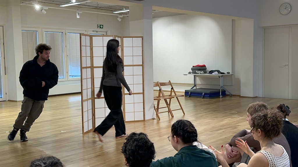 Two actors chase each other around the folding room divider prop in mid-performance of ''Faith Hope Charity'. A few students sit in the audience watching the performance.